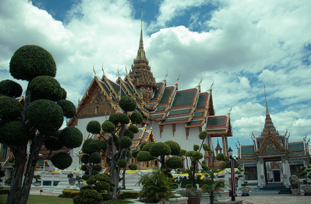 Exterior view of the ornate Thai architecture and spires of the Grand Palace under a partly cloudy sky in Bangkok.