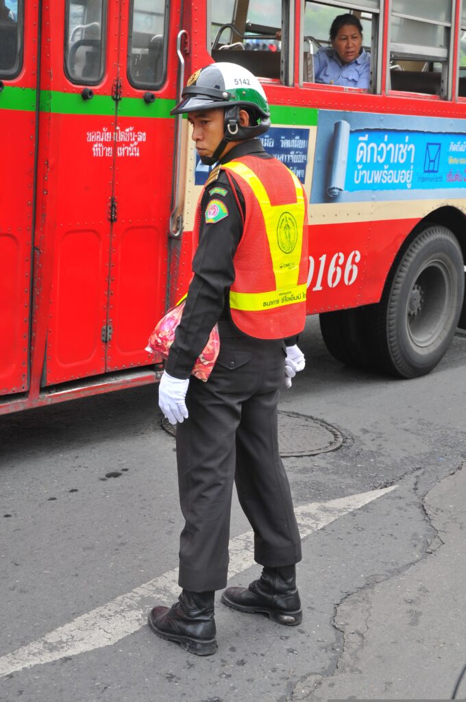 Royal Thai traffic police officer in uniform and helmet directing traffic next to a red BMTA public bus on a street in Bangkok Thailand.