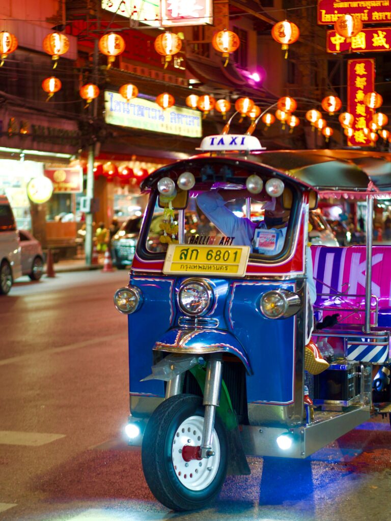 Illuminated motorized tuk-tuk waiting under red and orange hanging lanterns on a busy night street in Bangkok.
