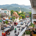 A digital nomad working on a laptop from a balcony cafe overlooking a busy chiang mai street with Tuk-tuks, Thai temples, and mountains in the background, illustrating the 2026 Thailand Digital Nomad Lifestyle.