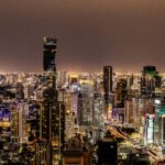 A panoramic nighttime aerial photograph of the Bangkok city skyline. The dense cityscape is a sea of illuminated windows and streetlights. The prominent King Power Mahanakhon skyscraper, with its distinctive spiral/pixelated glass facade, dominates the center-left. Other lit skyscrapers fill the background under a dark sky.