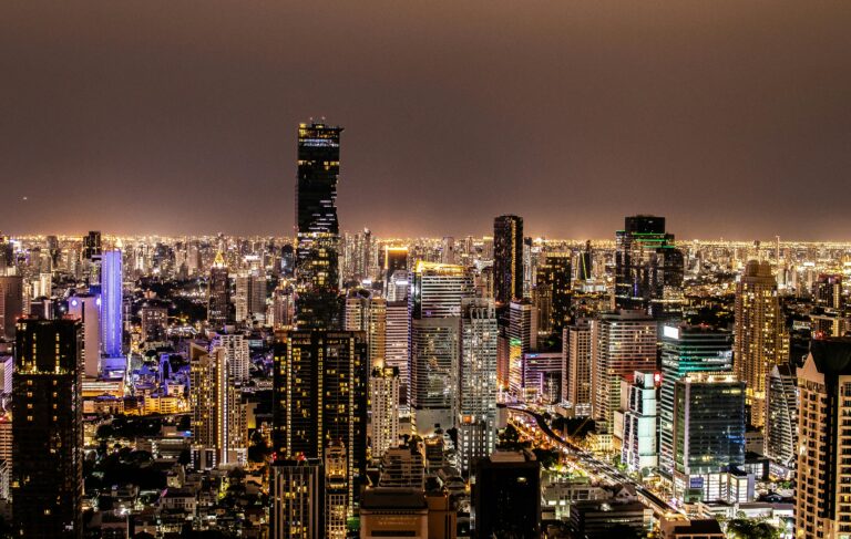 A panoramic nighttime aerial photograph of the Bangkok city skyline. The dense cityscape is a sea of illuminated windows and streetlights. The prominent King Power Mahanakhon skyscraper, with its distinctive spiral/pixelated glass facade, dominates the center-left. Other lit skyscrapers fill the background under a dark sky.