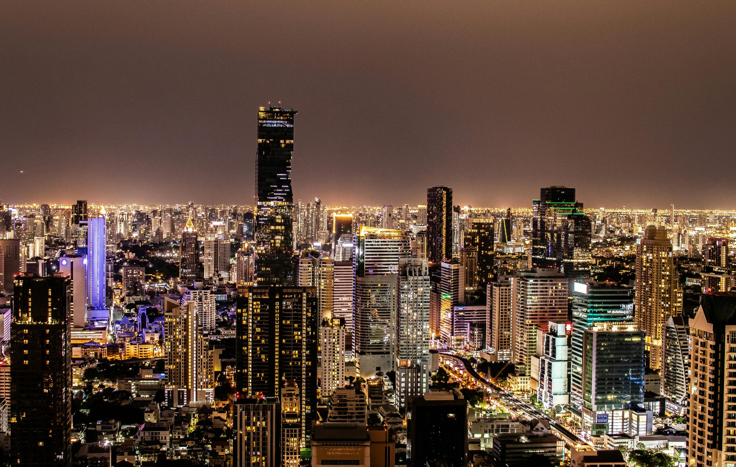 A panoramic nighttime aerial photograph of the Bangkok city skyline. The dense cityscape is a sea of illuminated windows and streetlights. The prominent King Power Mahanakhon skyscraper, with its distinctive spiral/pixelated glass facade, dominates the center-left. Other lit skyscrapers fill the background under a dark sky.