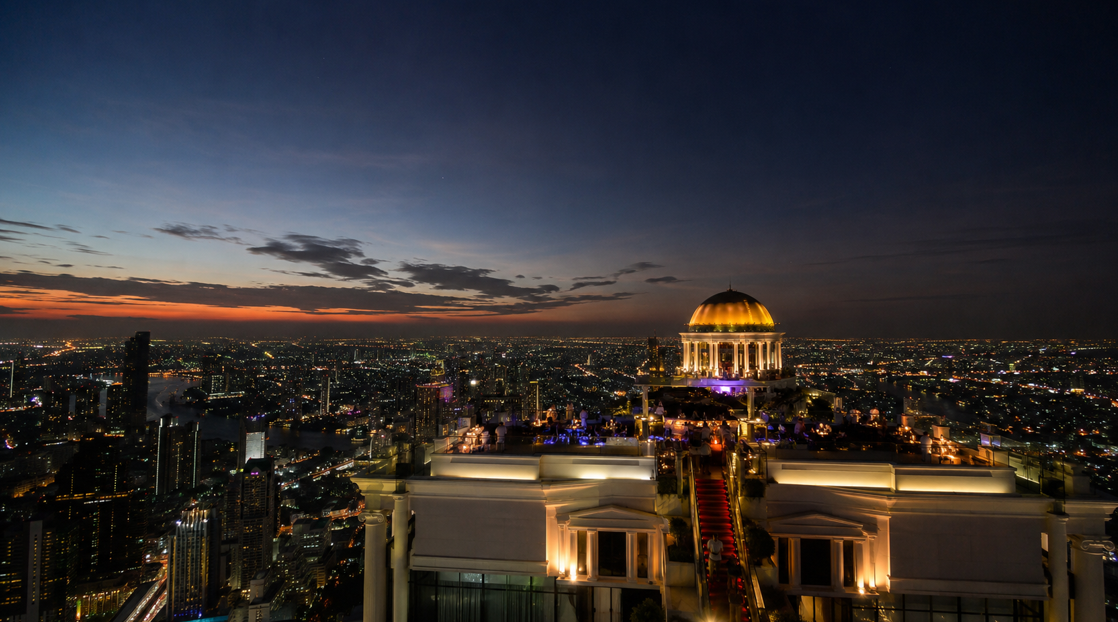 Wide angle evening to night view of Sky Bar at Lebua Bangkok rooftop bar with Bangkok skyline lights, luxury rooftop drinks, slow crowd atmosphere, and panoramic cityscape in Thailand.