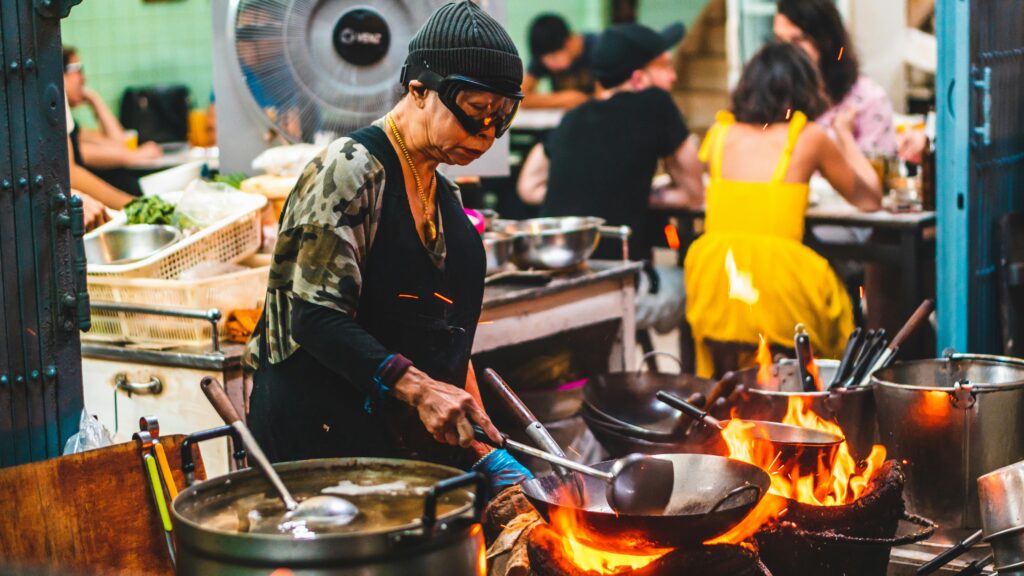 Michelin-starred chef Jay Fai (Supinya Junsuta) wearing her signature black beanie and ski goggles while cooking with a wok over a blazing charcoal fire in her Bangkok street-food restaurant.