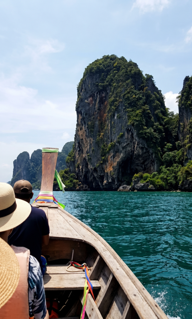 A first-person perspective photograph taken from the inside of a traditional Thai long-tail boat, showing the wooden hull and passengers in sun hats as they approach a massive, forest-covered limestone cliff rising from the turquoise sea.