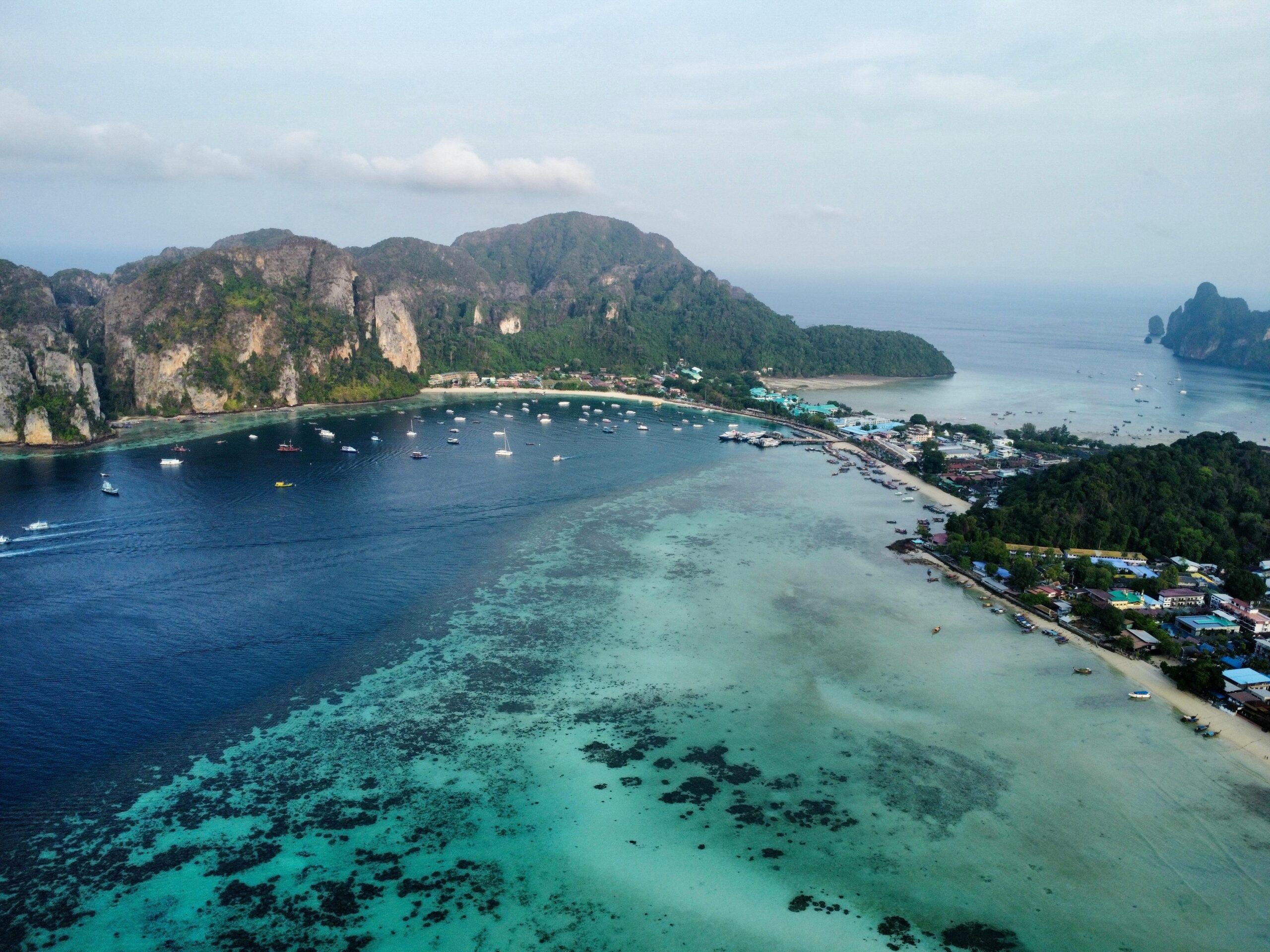 Aerial drone view of Ton Sai Bay and Loh Dalum Beach on Koh Phi Phi Don island in Thailand, showing the narrow sand isthmus, turquoise coral reefs, limestone cliffs, and boats moored in the deep blue bay.