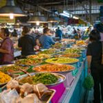 A bustling indoor Bangkok food market featuring a long counter of traditional Thai dishes in large metal platters with locals ordering street food.