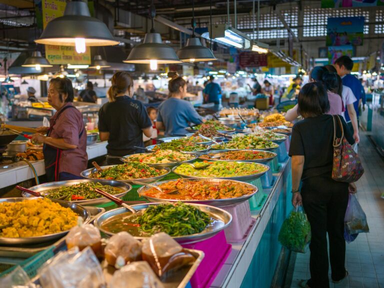 A bustling indoor Bangkok food market featuring a long counter of traditional Thai dishes in large metal platters with locals ordering street food.
