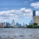Bangkok skyline viewed from the Chao Phraya River with skyscrapers and waterfront buildings under a blue cloudy sky.