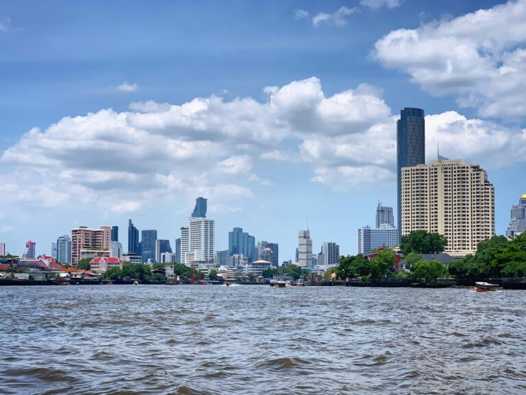 Bangkok skyline viewed from the Chao Phraya River with skyscrapers and waterfront buildings under a blue cloudy sky.