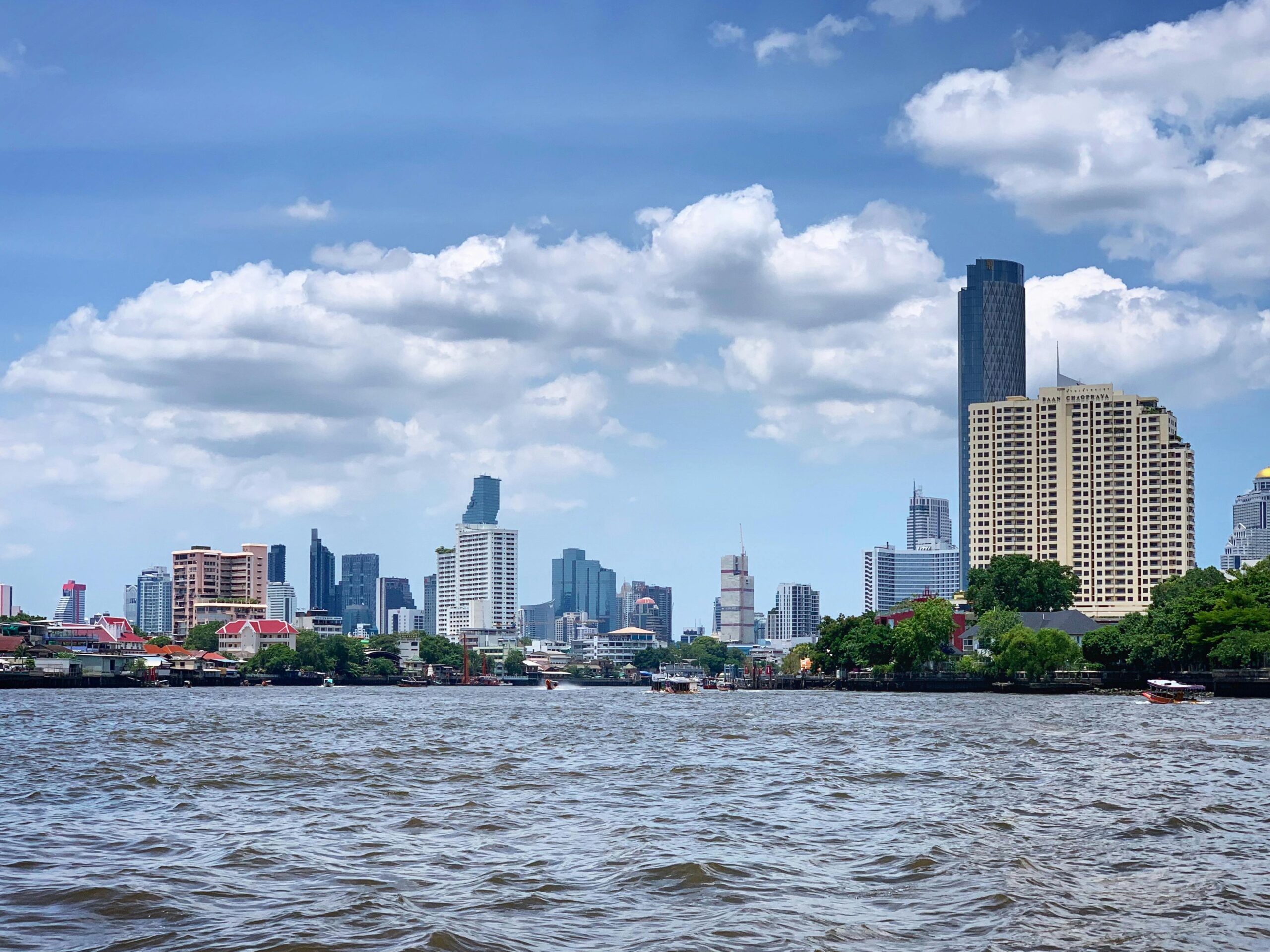 Bangkok skyline viewed from the Chao Phraya River with skyscrapers and waterfront buildings under a blue cloudy sky.