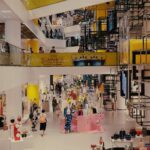 Wide-angle view of a multi-level modern shopping mall in Bangkok, Thailand, featuring glass balconies, trendy clothing displays, and geometric architectural installations.