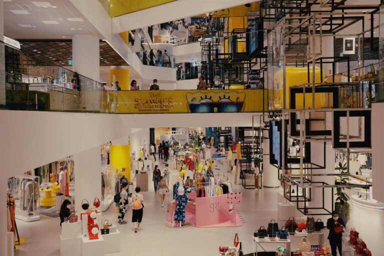 Wide-angle view of a multi-level modern shopping mall in Bangkok, Thailand, featuring glass balconies, trendy clothing displays, and geometric architectural installations.