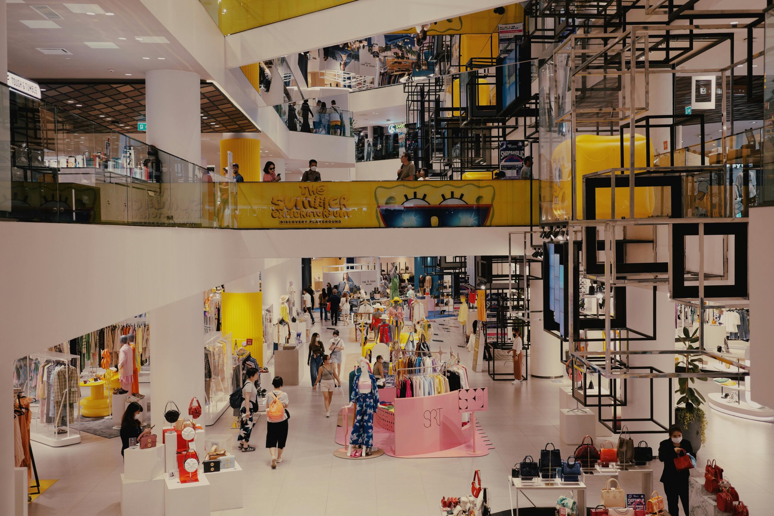 Wide-angle view of a multi-level modern shopping mall in Bangkok, Thailand, featuring glass balconies, trendy clothing displays, and geometric architectural installations.