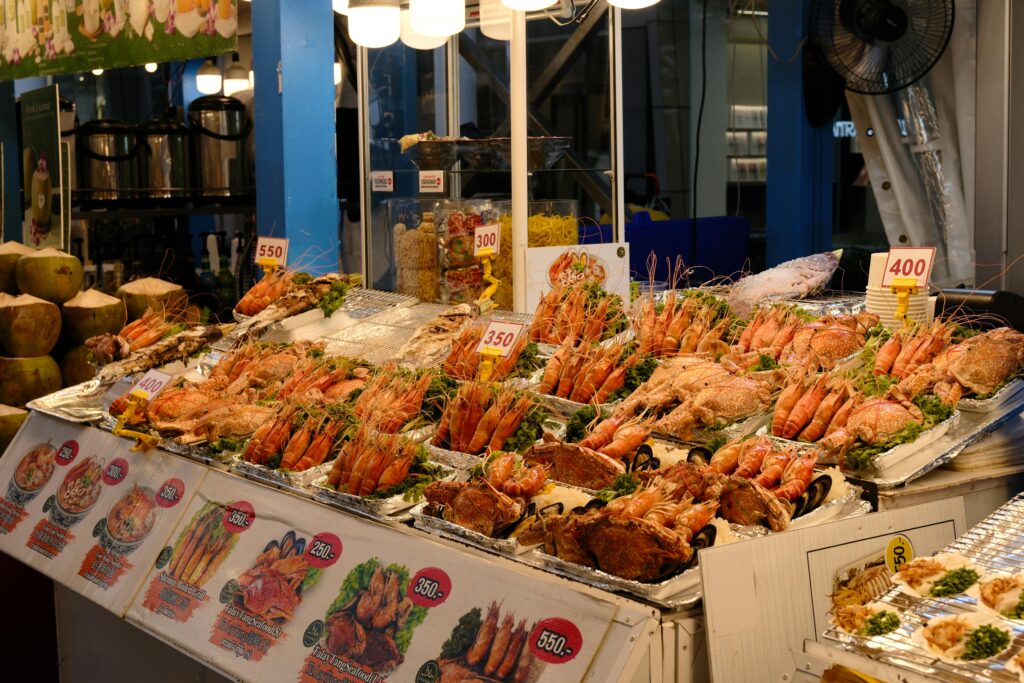 A brightly lit outdoor night market seafood stall in Thailand displaying a massive, vibrant arrangement of fresh prawns, crabs, whole fish, and shellfish on ice beds, complete with price tags and illustrated menu signs in the foreground.