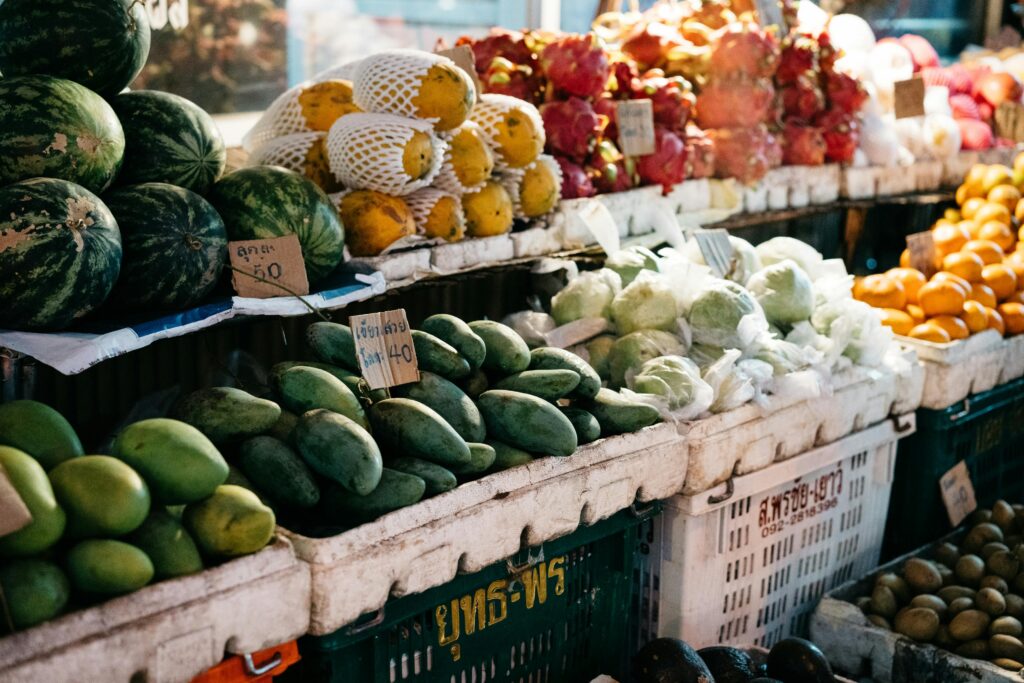 A colorful outdoor market stall in Thailand filled with rows of fresh tropical fruits, including whole watermelons, green mangoes with cardboard price tags in Thai script, papayas, dragon fruit, and citrus.