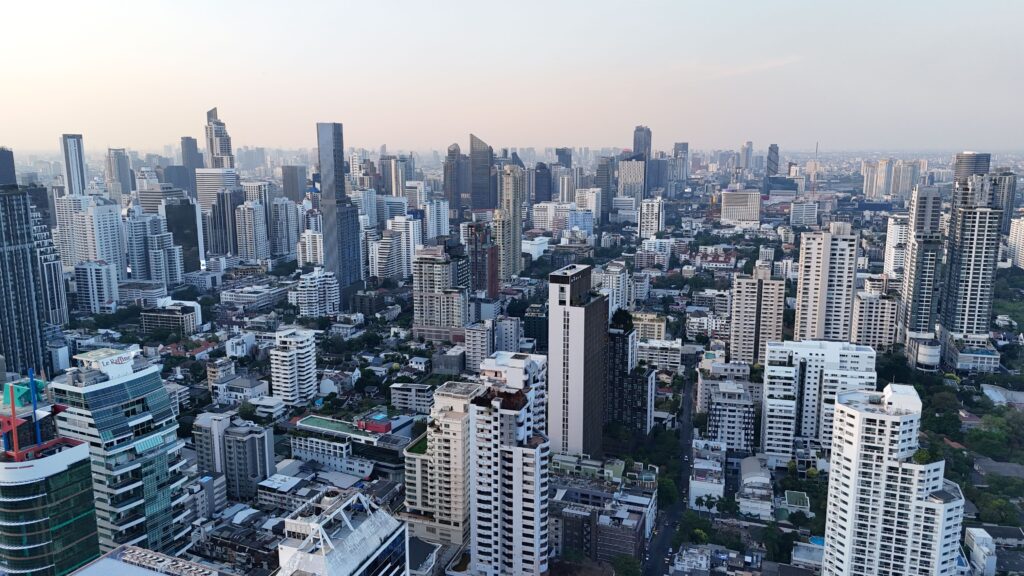 An aerial, wide-angle view of the dense Bangkok skyline at dusk, showcasing a vast sprawl of modern skyscrapers, high-rise residential buildings, and urban architecture stretching toward a hazy horizon under a soft evening sky.