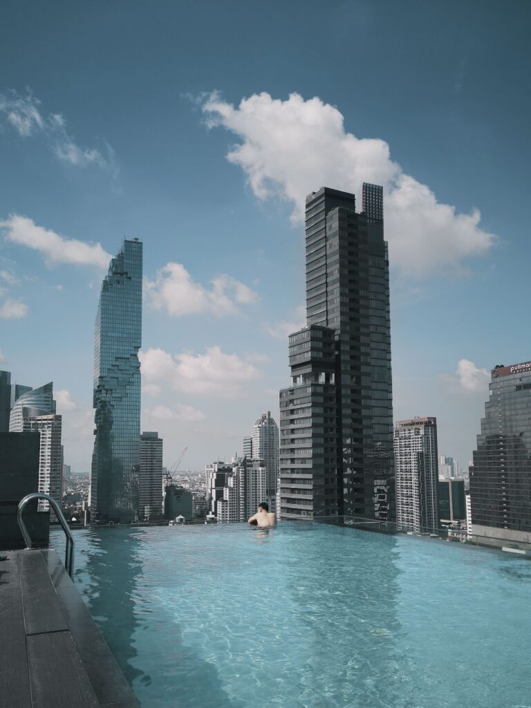 A breathtaking rooftop pool offers a stunning view of Bangkok's modern skyline under clear skies. A person is seen from behind, relaxing at the edge of a crystal-clear, blue infinity pool on a high-rise rooftop. In the background, prominent skyscrapers tower into the air, including the iconic, pixelated glass structure of King Power Mahanakhon on the left and a tall dark skyscraper on the right, all set against a bright blue sky with scattered white clouds.