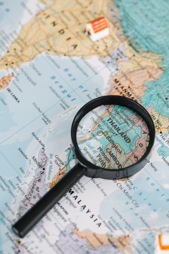 A black-framed magnifying glass rests on a detailed world map, enlarging Thailand and Bangkok, with a small decorative house figurine placed near the Bay of Bengal.