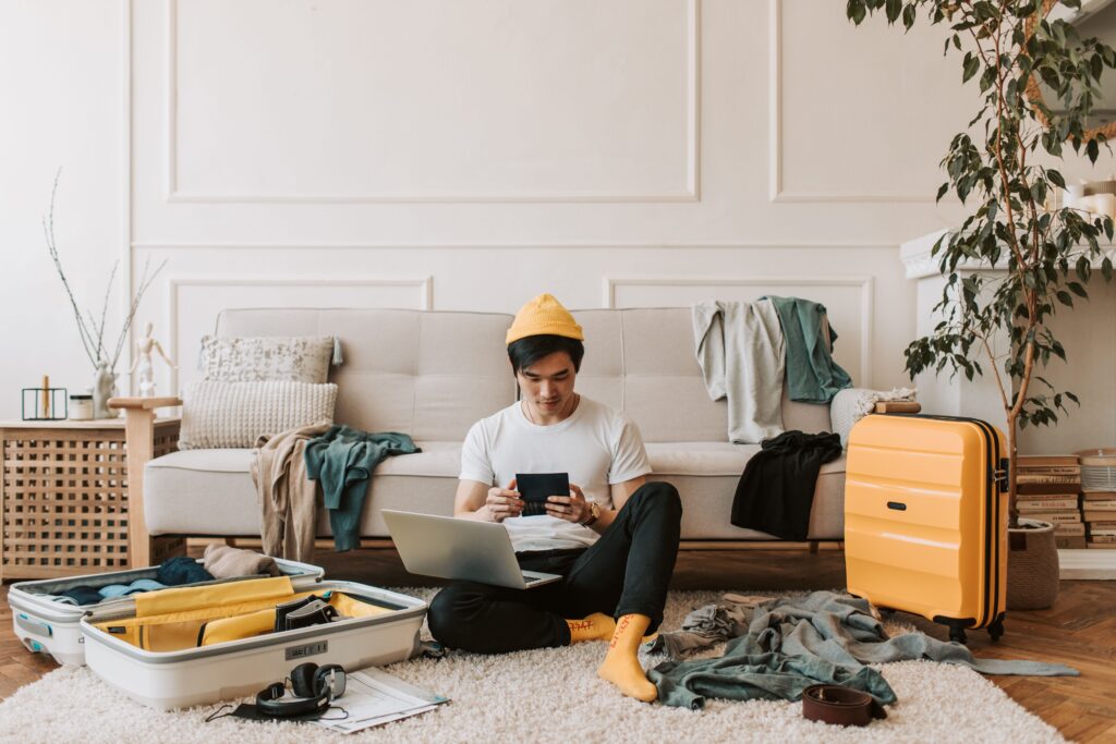 A young man wearing a yellow beanie, white t-shirt, dark trousers, and bright yellow socks sits cross-legged on a cream-colored rug in a brightly lit living room. He holds a dark smartphone in both hands while balancing an open silver laptop on his lap. Beside him on the floor is an open white suitcase packed with clothes, pair of headphones, and travel papers, while a vibrant yellow hard-shell suitcase stands upright to his right. In the background, clothes are draped over a light beige sofa near a tall indoor potted plant.