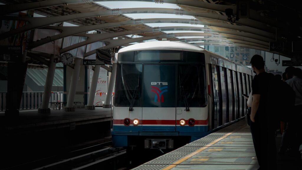 A Bangkok BTS Skytrain with its headlights on arrives at an elevated, covered urban metro station platform. Commuters are standing on the platform edge, waiting to board the white and blue train under a modern arched roof structure.