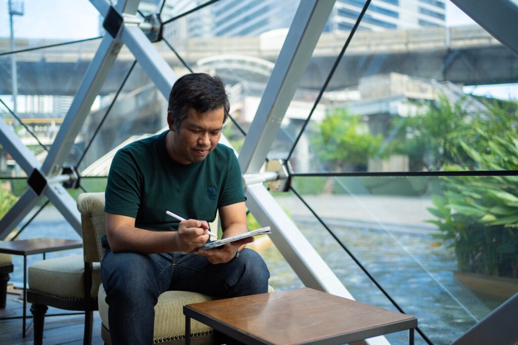 A man wearing a dark green t-shirt and jeans sits in a modern cafe with geometric structural framing, using a stylus on a tablet.