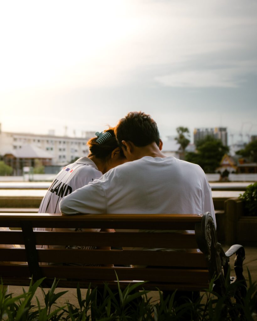 A view from behind of a couple sitting close together on a wooden park bench during a peaceful sunset. The man wears a white long-sleeve shirt and has his arm wrapped around the woman, who is wearing a white jersey with "THAILAND" printed on the back. They lean their heads together affectionately, with green foliage in the foreground and a soft city horizon under the glowing evening sky.