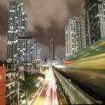 Bangkok Digital Nomads Guide and a long exposure night photograph in Bangkok showing a blurred BTS Skytrain speeding past on an elevated track on the right, leaving streaks of green and yellow light. Below, a busy street is filled with bright white and red car light trails. High-rise residential and commercial buildings with illuminated windows stand under a cloudy night sky in the background.