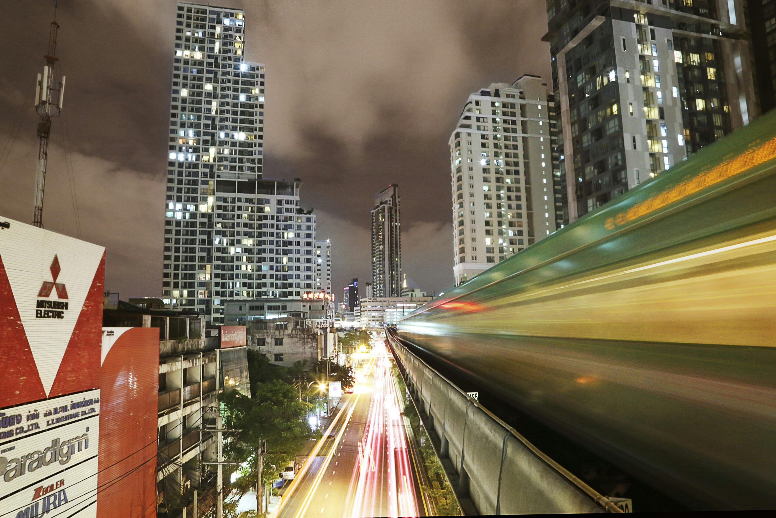 Bangkok Digital Nomads Guide and a long exposure night photograph in Bangkok showing a blurred BTS Skytrain speeding past on an elevated track on the right, leaving streaks of green and yellow light. Below, a busy street is filled with bright white and red car light trails. High-rise residential and commercial buildings with illuminated windows stand under a cloudy night sky in the background.