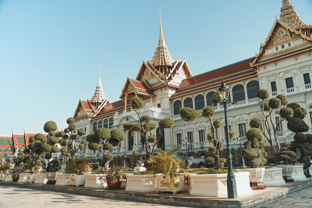 The grand Chakri Maha Prasat Throne Hall inside the Grand Palace complex in Bangkok, Thailand. The landmark building features a unique blend of European Renaissance architecture on its lower structure combined with traditional tiered Thai roofs and gilded spires, fronted by a manicured courtyard with topiary trees under a clear blue sky.