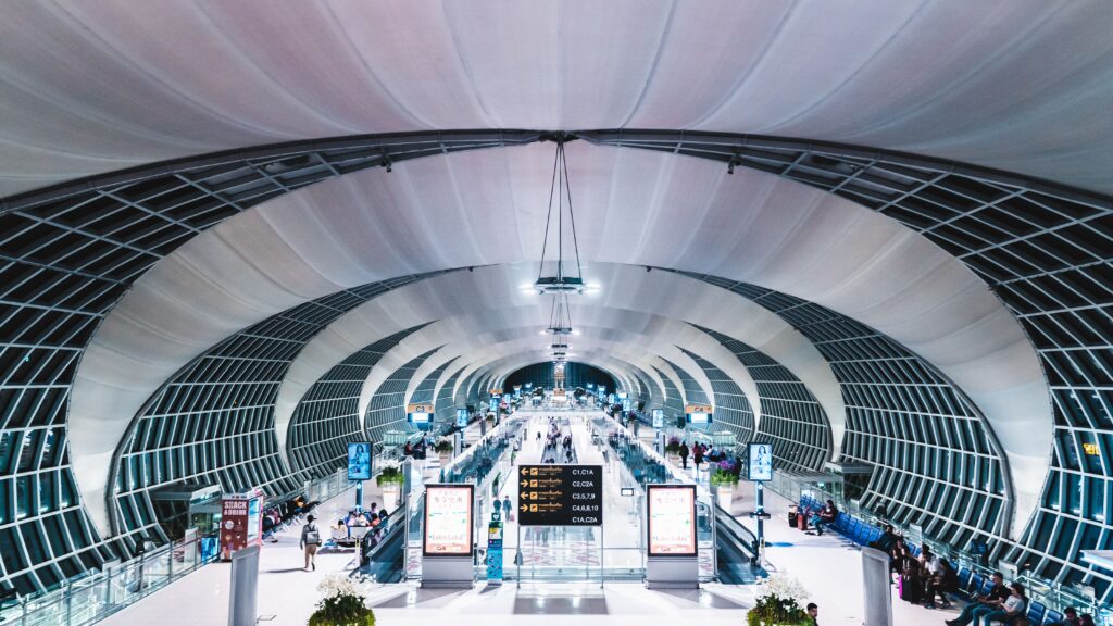 Symmetrical, wide-angle view of the futuristic, vaulted fabric roof and steel lattice structure of Suvarnabhumi Airport terminal in Bangkok, Thailand, with travelers walking below.