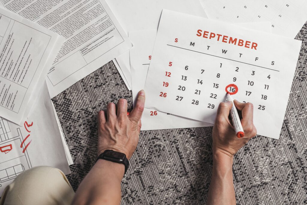 A person sits on a patterned carpet surrounded by travel documents and papers, using a red marker to circle dates on a September calendar page.