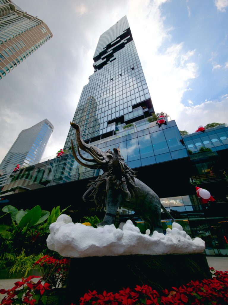 A low-angle shot of the pixelated King Power MahaNakhon skyscraper in Bangkok rising into a cloudy sky, with a large bronze statue of a three-headed elephant on a white cloud base surrounded by red flowers in the foreground.