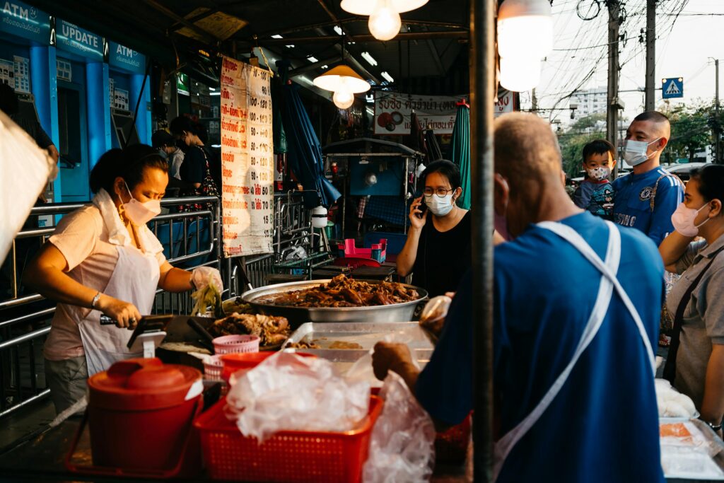 A female Thai street food vendor wearing a face mask selling food at her outdoor stall under hanging lights, surrounded by customers waiting for local dishes.