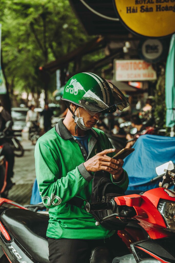 A delivery rider wearing a green Grab jacket and matching helmet checks a smartphone while sitting on a red motorbike parked along a busy street.