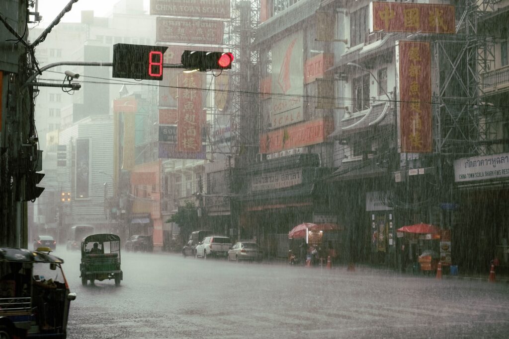 A wide street view of Bangkok's Chinatown during a heavy rainstorm. A red traffic light with an 8-second countdown timer hangs over the wet road. Below, a green tuk-tuk drives through the deluge alongside cars and roadside stalls shielded by red umbrellas. The multi-story buildings on the right are densely covered in large, traditional signs featuring Chinese characters.