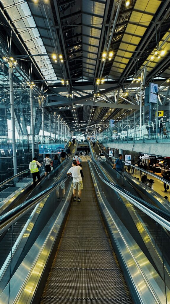 A vertical shot inside a massive, modern airport terminal showing travelers riding a long, central moving walkway. The interior features a soaring, industrial-style ceiling with an exposed steel truss structure, extensive glass paneled walls, and bright overhead lighting illuminating the concourse.