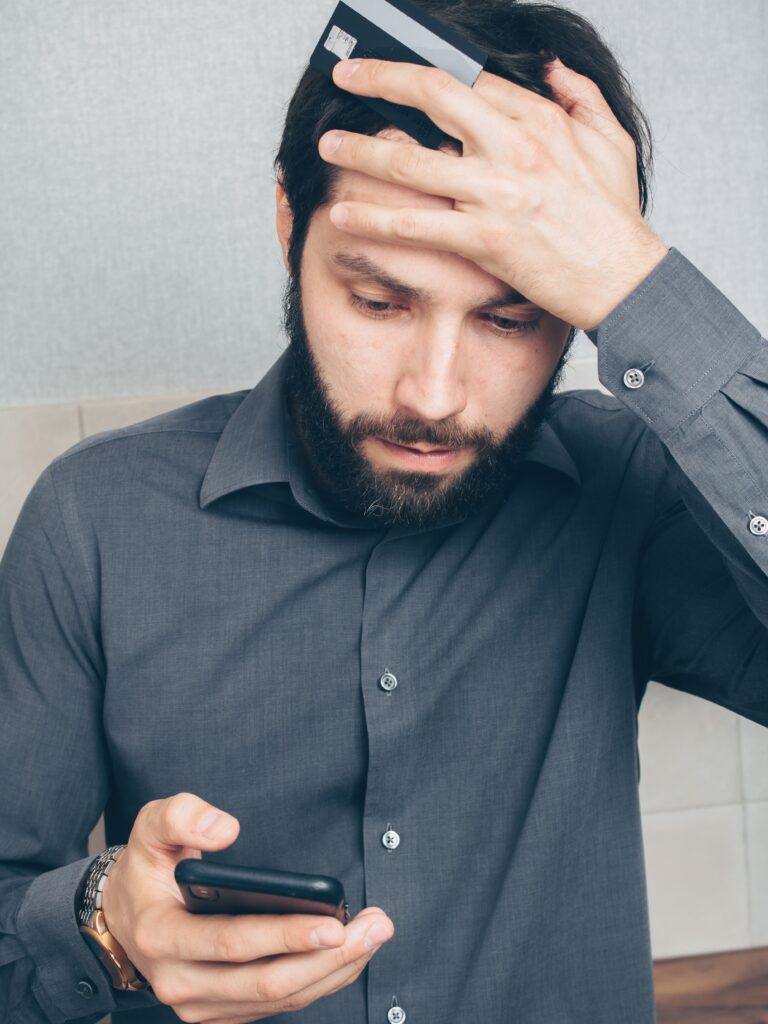 A bearded man wearing a dark grey button-down shirt looks down stressed and concerned while holding a credit card against his forehead with one hand and typing on a smartphone with the other.