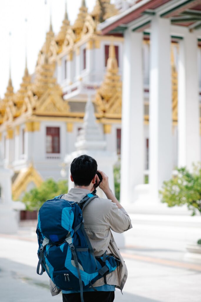 A view from behind of a solo traveler wearing a large blue backpack and a light brown shirt, holding a camera up to take a picture. In the softly blurred background stands a magnificent traditional Thai temple featuring white pillars and striking multi-tiered golden spires under bright daylight.