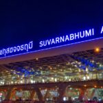 A wide-angle night photograph of the exterior of Suvarnabhumi Airport in Bangkok, Thailand, in this Bangkok Airport Survival Guide, featuring a massive, brightly illuminated blue neon sign displaying the airport's name in Thai script and English above a modern, multi-level glass and steel terminal structure.