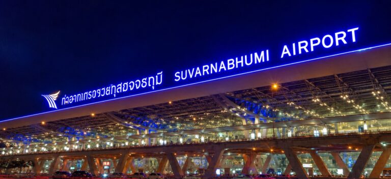 A wide-angle night photograph of the exterior of Suvarnabhumi Airport in Bangkok, Thailand, in this Bangkok Airport Survival Guide, featuring a massive, brightly illuminated blue neon sign displaying the airport's name in Thai script and English above a modern, multi-level glass and steel terminal structure.