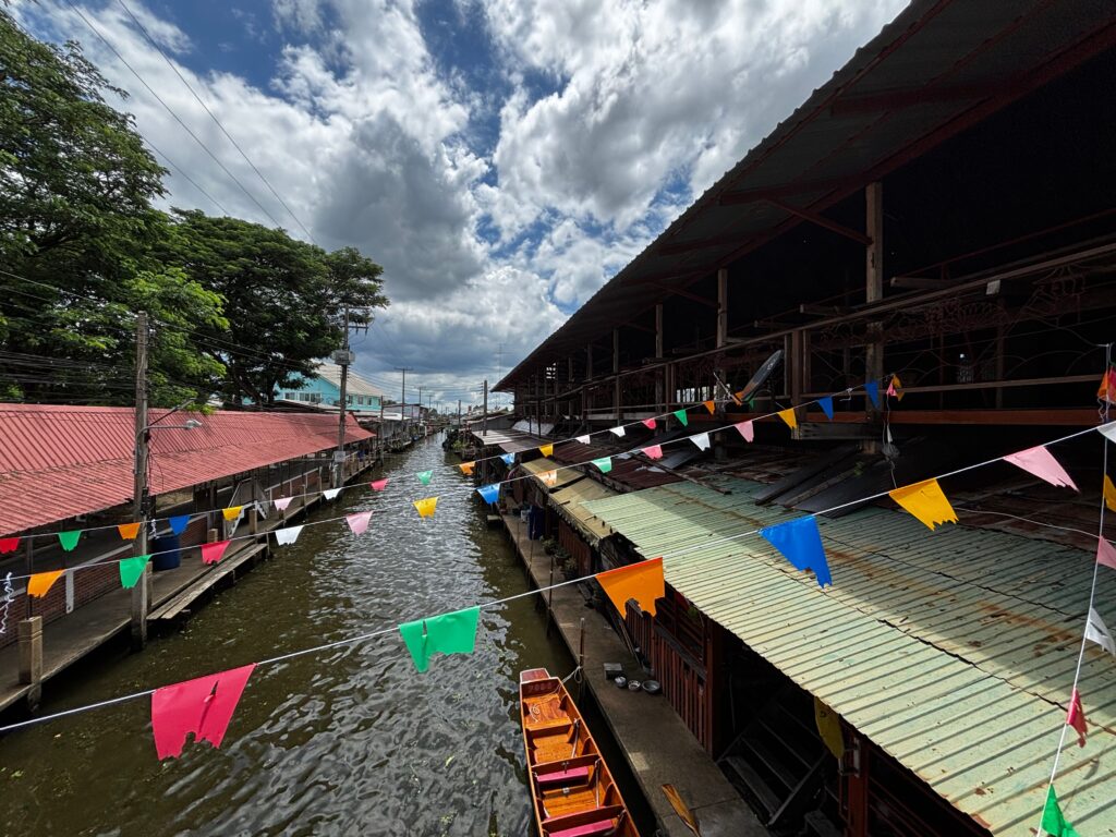 Amphawa Floating Marketing is one of the most popular weekend Bangkok day trips, in Thailand. This is a view down the canal at Amphawa Floating Market near Bangkok, featuring traditional wooden waterside shops, colorful bunting flags, and a docked longtail boat under a cloudy sky with historic wooden homes.