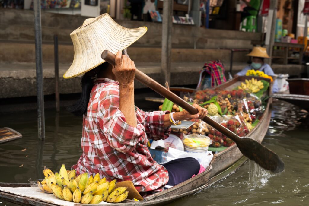 A local vendor wearing a traditional woven hat and a red plaid shirt paddles a wooden boat filled with fresh bananas, rambutans, and tropical fruits along a canal at a Bangkok floating market.
