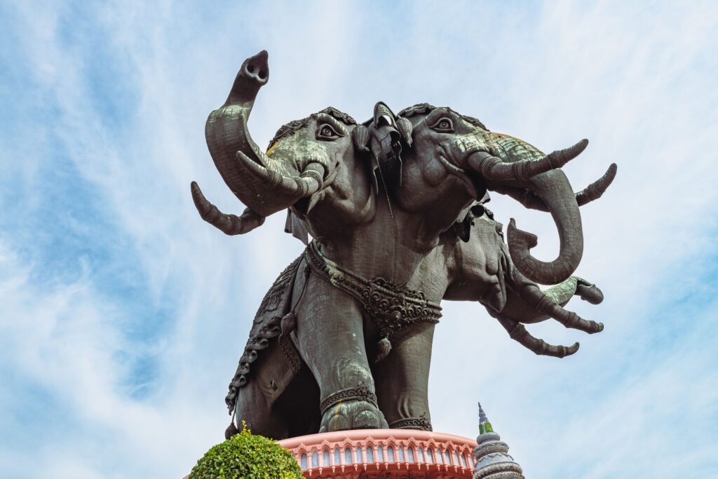 A massive, intricately detailed three-headed bronze elephant statue towering against a bright blue sky at the Erawan Museum, an iconic Bangkok day trip in Samut Prakan, Thailand.