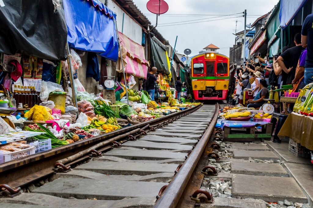 A bright yellow and red train passing inches from vendor stalls selling fresh produce on the tracks at Mae Klong Railway Market, one of the most popular Bangkok Day Trips in Samut Songkhram, Thailand.