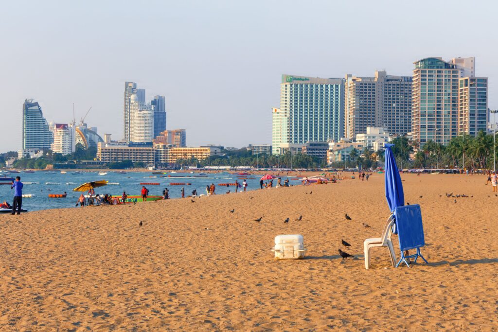 A wide view of Pattaya Beach featuring golden sand, tourists enjoying the water, and a modern city skyline of resorts, highlighting a popular coastal day trip from Bangkok, Thailand.