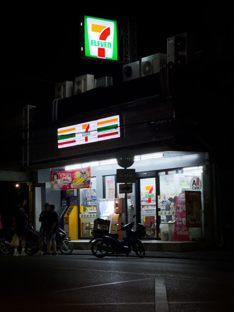 A brightly lit 7-Eleven convenience store glowing on a dark street at night in Thailand, with motorbikes parked out front and pedestrians standing near the entrance.