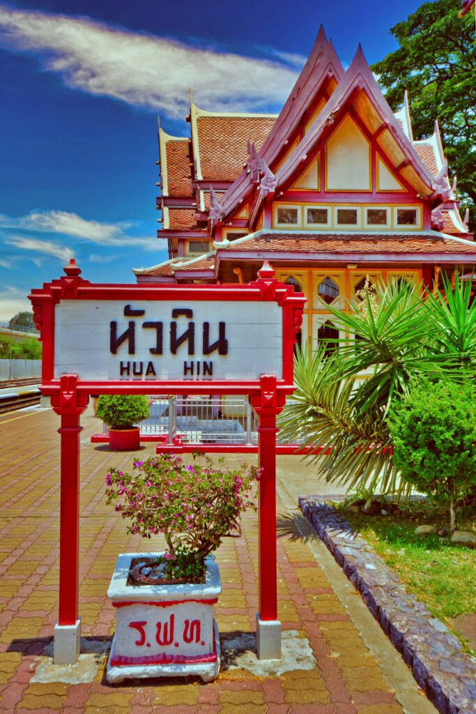 The iconic red and white wooden Hua Hin Railway Station sign standing in front of the traditional Thai-style Royal Waiting Room, a historic and photogenic Bangkok day trip destination.